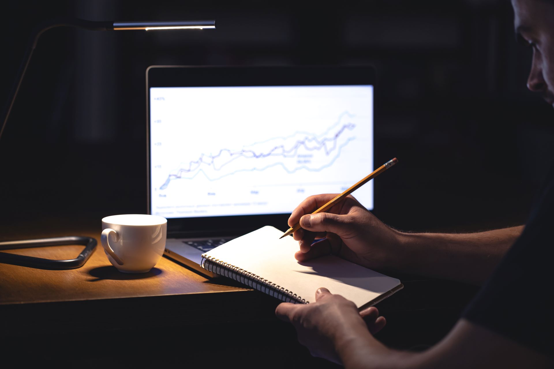 man-sits-front-laptop-with-chart-notepad-night